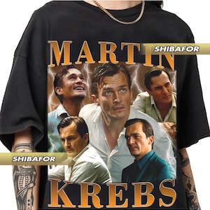 Könnte beinhalten: Schwarzes T-Shirt mit einem Retro-Design, das das Bild eines Mannes und den Text "MARTIN KREBS" in Orange zeigt. Die Grafik enthält mehrere Bilder des Mannes und Blitzakzente. Das T-Shirt hat einen klassischen Rundhalsausschnitt.