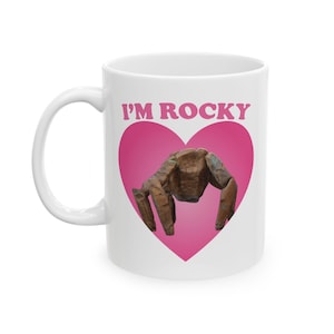 May include: White ceramic mug featuring a pink heart design with the text "I'M ROCKY" in pink. The heart contains a brown, textured sculpture. The mug has a white handle.