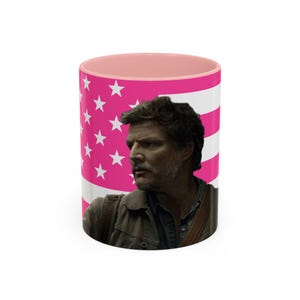 May include: A white ceramic coffee mug with a pink interior and a vibrant pink and white American flag design. The mug features a portrait of a man on the front.