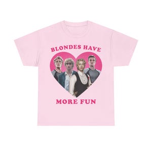 Puede incluir: Camiseta rosa claro con un diseño en forma de corazón rosa. El corazón contiene cuatro hombres y el texto "BLONDES HAVE MORE FUN". La camiseta es de material suave.