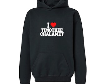 Może przedstawiać: Czarna bluza z kapturem z przednią kieszenią i napisem "I ❤️ Timothée Chalamet" w kolorze białym z czerwonym sercem. Bluza ma kaptur ze sznurkiem i długie rękawy.