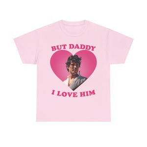 Puede incluir: Camiseta rosa con un gráfico en forma de corazón que presenta la cara de un hombre y el texto "But Daddy I Love Him".