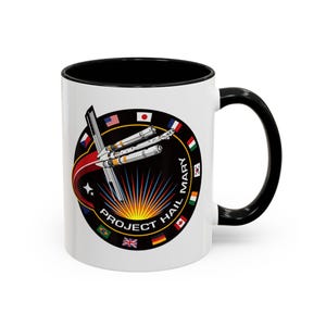 Könnte beinhalten: Weiße Keramik-Tasse mit schwarzem Innenraum und Griff. Die Tasse zeigt ein rundes Emblem mit Raketendesign, dem Text "PROJECT HAIL MARY" und Flaggen verschiedener Länder. Das Design beinhaltet einen roten und orangenen Ausbruch.