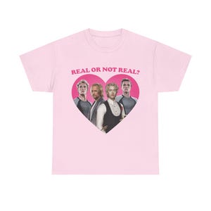 Può includere: Una t-shirt a maniche corte color rosa chiaro con una grafica centrale a forma di cuore rosa contenente quattro figure maschili. Sopra il cuore, il testo REAL OR NOT REAL? è stampato in lettere maiuscole rosa. Girocollo e maniche corte.