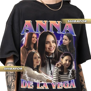 May include: Black t-shirt featuring a graphic of a woman with the name "ANNA DE LA VEGA" in a retro style. The design includes multiple images of the woman and lightning bolt accents, with the brand name "SHIBAFOR" visible.