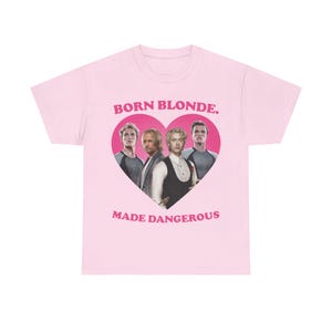 May include: A light pink short-sleeved t-shirt featuring a large dark pink heart graphic containing four men. Above the heart is the text BORN BLONDE. and below the heart is the text MADE DANGEROUS. The shirt has a crew neckline and a standard fit.