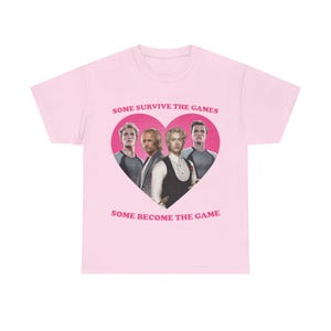 Puede incluir: Una camiseta de manga corta de color rosa claro con un gráfico central de un corazón rosa oscuro que contiene cuatro figuras masculinas. El texto "SOME SURVIVE THE GAMES" aparece sobre el corazón y "SOME BECOME THE GAME" debajo.