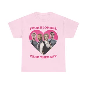 Può includere: Una t-shirt a maniche corte rosa chiaro con una grande grafica a forma di cuore rosa scuro sul petto. All'interno del cuore ci sono quattro uomini biondi, con il testo "FOUR BLONDES, ZERO THERAPY" in lettere maiuscole rosa scuro.
