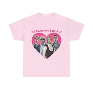 Può includere: Una t-shirt a maniche corte rosa chiaro con una grafica centrale a forma di cuore rosa scuro contenente quattro ritratti di un uomo con i capelli biondi. Sopra il cuore, il testo REAL OR NOT REAL? è stampato in un carattere sans-serif.