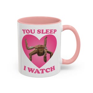 May include: White ceramic mug with a pink interior and handle. The mug features the text "YOU SLEEP I WATCH" in pink, with a brown figure inside a pink heart.