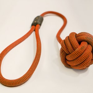 Op de afbeelding: Oranje en groen touwspeelgoed voor honden met een geknoopte bal aan het ene uiteinde en een lus aan het andere uiteinde.