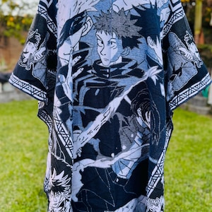 Puede incluir: Un poncho con capucha con un estampado gráfico de estilo anime en negro, blanco y azul. El diseño incluye múltiples personajes y patrones geométricos. El poncho tiene un dobladillo con flecos y está hecho de una tela suave.