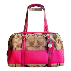 May include: A beige and brown monogram patterned handbag with vibrant hot pink leather trim and handles. The bag features silver-toned hardware, including circular rings, rivets, and a small circular hanging charm attached to the front strap.