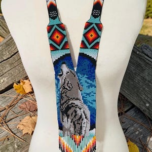 May include: A beaded necklace featuring a central pendant depicting a howling wolf against a blue background. The design includes geometric patterns in turquoise, black, red, orange, and white beads, finished with a multi-colored beaded fringe.