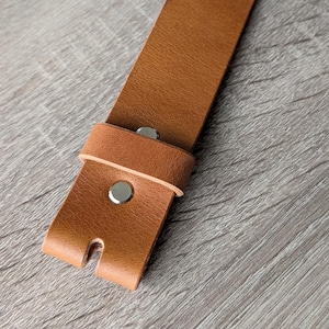 May include: A tan leather strap featuring a smooth texture and a rectangular end with a small vertical slit. The strap is secured by a matching leather loop and two polished silver-toned metal rivets positioned along the centre of the material.