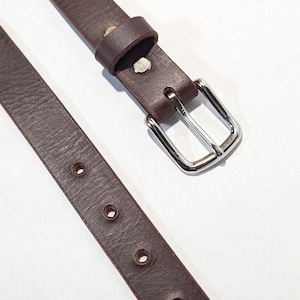 May include: A narrow dark brown leather belt featuring a textured surface, a single silver-toned metal pin buckle, a matching leather keeper loop, and a series of evenly spaced circular holes punched along the length of the strap.