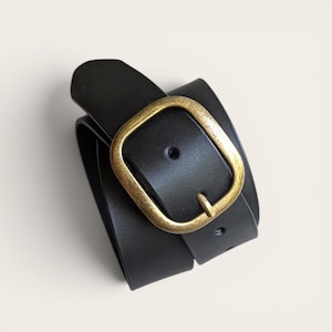 Peut inclure: Une ceinture noire avec une finition lisse et mate et une boucle arrondie en laiton de style antique. La ceinture est enroul&eacute;e, montrant une ardillon unique et une s&eacute;rie de trous de r&eacute;glage le long de la sangle, avec une largeur uniforme.