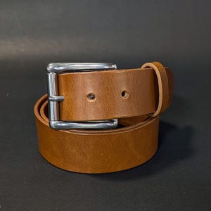 May include: A tan leather belt featuring a smooth finish and a rectangular silver-toned metal buckle. The belt is coiled into a circular shape and includes a single leather keeper loop near the buckle, with two visible holes for adjustment.