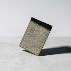 May include: A rectangular card holder made of textured, olive-green leather. The accessory features two vertical card slots on the front surface, with visible stitching along the edges and a consistent grain pattern across the entire exterior surface.