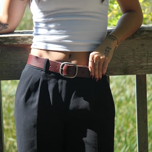 Peut inclure: Une ceinture en cuir marron fonc&eacute; avec une finition lisse et une boucle m&eacute;tallique rectangulaire arrondie. La ceinture est pass&eacute;e dans les passants d'un pantalon noir, assurant un maintien ajust&eacute; autour de la taille. Le design est simple.