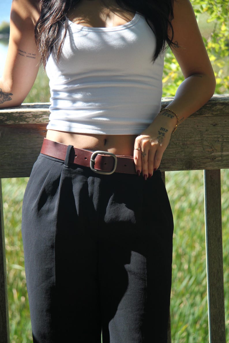 Peut inclure: Une ceinture en cuir marron fonc&eacute; avec une finition lisse et une boucle m&eacute;tallique rectangulaire arrondie. La ceinture est pass&eacute;e dans les passants d'un pantalon noir, assurant un maintien ajust&eacute; autour de la taille. Le design est simple.