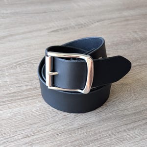 May include: A black belt coiled into a circular shape, featuring a smooth matte finish and a polished silver-toned metal buckle with a single prong. The belt strap has a pointed end and is constructed from a solid, uniform material throughout.