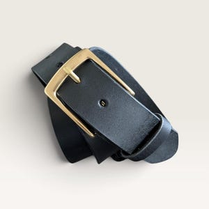 May include: A black belt featuring a smooth, matte finish and a rectangular gold-toned metal buckle. The belt strap is threaded through the buckle, which includes a single prong and a matching black leather keeper loop to secure the end of the strap.