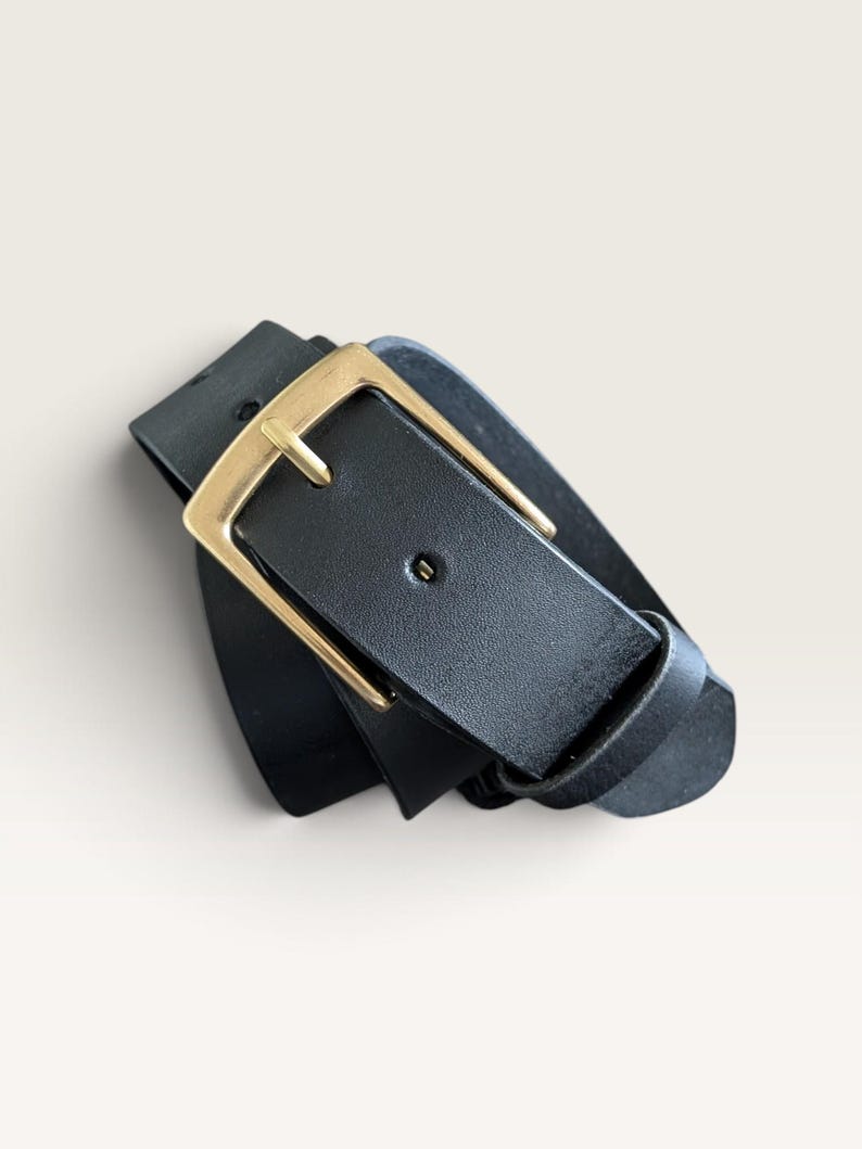 May include: A black belt featuring a smooth, matte finish and a rectangular gold-toned metal buckle. The belt strap is threaded through the buckle, which includes a single prong and a matching black leather keeper loop to secure the end of the strap.