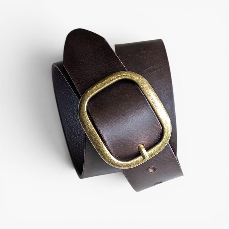 Peut inclure: Une ceinture en cuir marron fonc&eacute; avec une finition lisse et mate, dot&eacute;e d'une boucle en m&eacute;tal de couleur laiton antique aux bords arrondis. La ceinture est enroul&eacute;e, r&eacute;v&eacute;lant la texture du cuir et l'&eacute;clat m&eacute;tallique de la boucle.