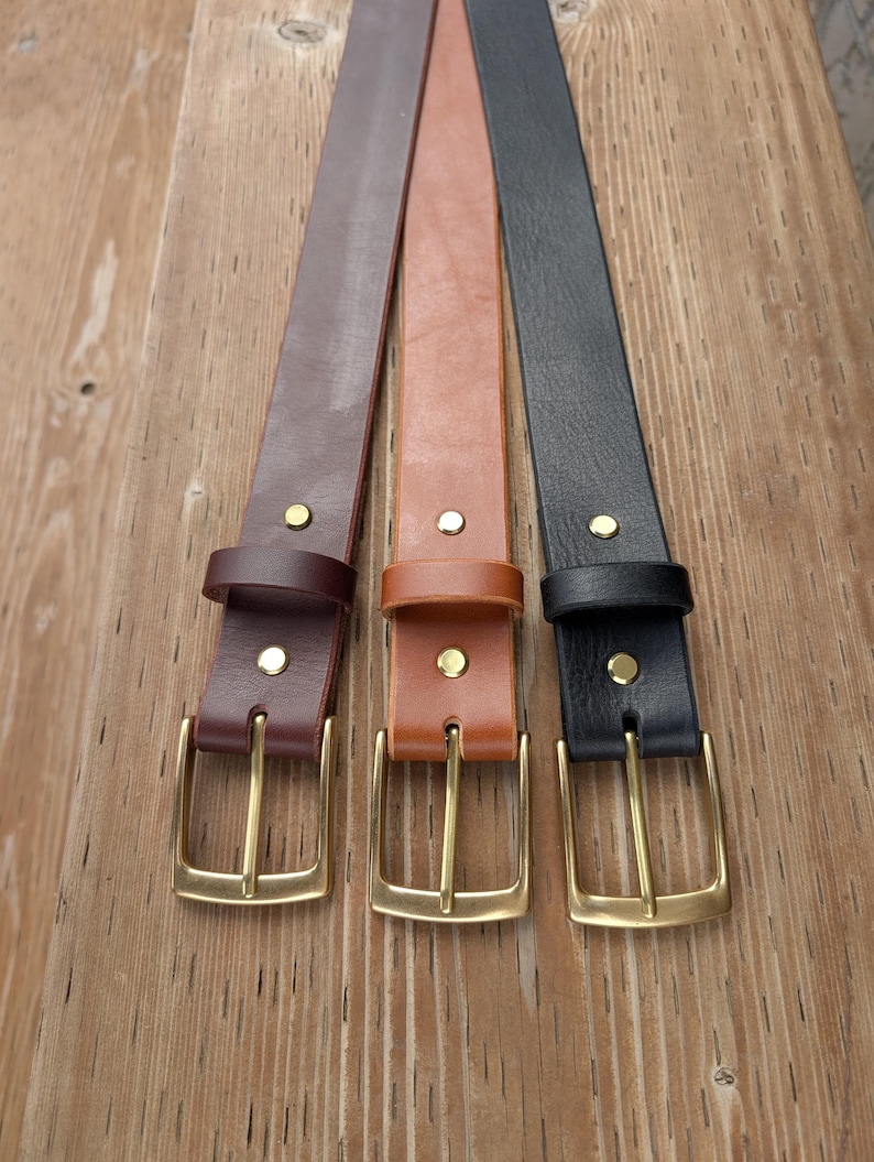 May include: Three leather belts in dark brown, tan, and black. Each belt features a smooth finish, a matching leather keeper loop, two gold-toned metal rivets, and a rectangular gold-toned metal buckle with a single prong for secure fastening.