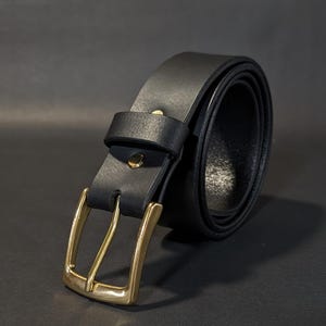 May include: A black belt featuring a smooth matte finish and a polished brass-toned metal buckle. The belt includes a matching black leather keeper loop and a single brass-toned rivet securing the strap near the buckle assembly.