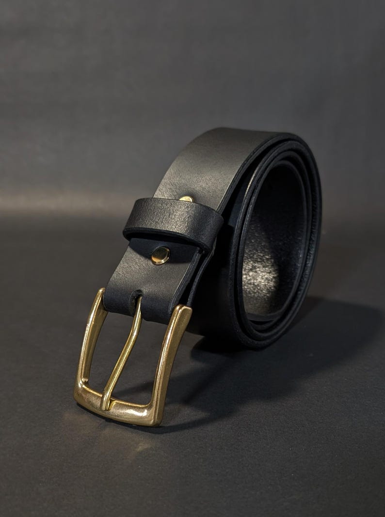 May include: A black belt featuring a smooth matte finish and a polished brass-toned metal buckle. The belt includes a matching black leather keeper loop and a single brass-toned rivet securing the strap near the buckle assembly.