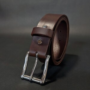 May include: A dark brown belt featuring a smooth finish and a rectangular silver-toned metal buckle. The belt includes a matching leather keeper loop and a single silver-toned metal rivet securing the strap near the buckle assembly.