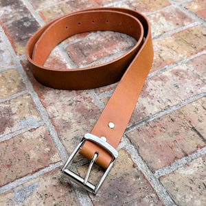 May include: A tan leather belt featuring a smooth finish and a rectangular silver-toned metal buckle. The belt strap includes a series of punched holes for adjustment and is secured with two small silver-toned metal rivets near the buckle.