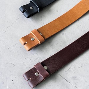 May include: Three leather belts in black, tan, and dark brown. Each belt features a smooth texture, a single leather keeper loop, and a silver-toned metal rivet securing the strap end. The belts are arranged diagonally across the frame.