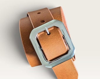 Peut inclure: Une ceinture en cuir beige dotée d'une boucle rectangulaire en métal argenté poli. La sangle de la ceinture présente une texture grainée visible et deux trous pré-percés près de la boucle, offrant un design classique avec une finition métallique.
