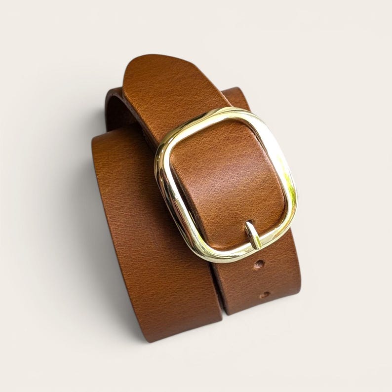 Peut inclure: Une ceinture en cuir marron clair dot&eacute;e d'une boucle en m&eacute;tal dor&eacute; de forme rectangulaire arrondie et lisse. La sangle pr&eacute;sente une texture grain&eacute;e visible et est enroul&eacute;e, avec une ardillon ins&eacute;r&eacute; dans l'un des trous pr&eacute;-perfor&eacute;s.