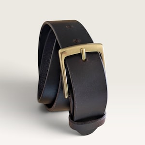 May include: A dark brown leather belt featuring a smooth finish and a rectangular brass-toned metal buckle. The belt is coiled, showing a single prong and a matching leather keeper loop near the buckle, with visible stitching along the edges.