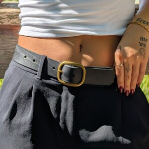 Peut inclure: Une ceinture en cuir noir avec une texture lisse et une boucle en m&eacute;tal de couleur laiton antique arrondie. La ceinture est pass&eacute;e dans les passants d'un pantalon noir, fix&eacute;e &agrave; la taille et port&eacute;e avec un haut blanc c&ocirc;tel&eacute;.