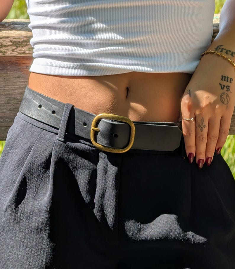 Peut inclure: Une ceinture en cuir noir avec une texture lisse et une boucle en m&eacute;tal de couleur laiton antique arrondie. La ceinture est pass&eacute;e dans les passants d'un pantalon noir, fix&eacute;e &agrave; la taille et port&eacute;e avec un haut blanc c&ocirc;tel&eacute;.