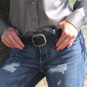 Peut inclure: Une chemise grise &agrave; boutons avec des manches longues, associ&eacute;e &agrave; un jean bleu en denim pr&eacute;sentant des zones us&eacute;es et des bords effiloch&eacute;s. Une ceinture en cuir noir avec une boucle en m&eacute;tal argent&eacute; mat et arrondi est port&eacute;e &agrave; la taille.