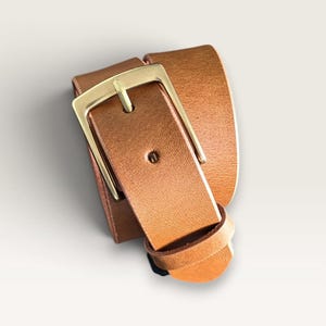 May include: A tan leather belt coiled into a loop, featuring a smooth texture and a rectangular gold-toned metal buckle. The belt strap is secured by a matching leather keeper loop, showcasing a clean design with visible stitching details.