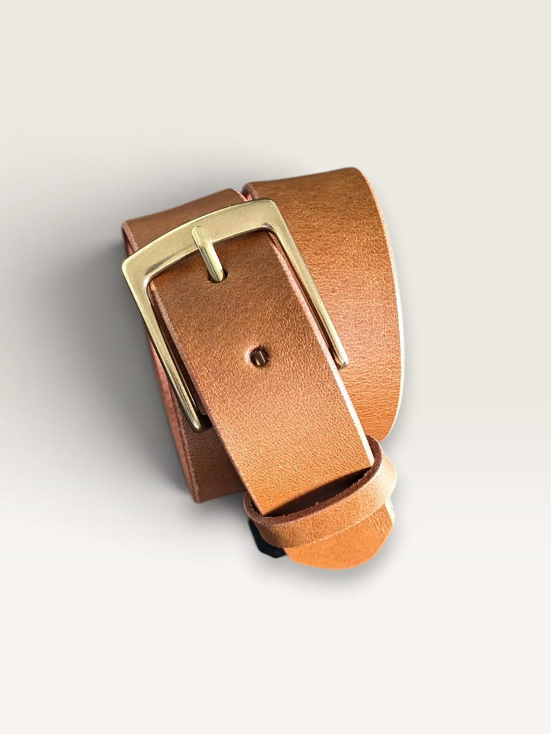 May include: A tan leather belt coiled into a loop, featuring a smooth texture and a rectangular gold-toned metal buckle. The belt strap is secured by a matching leather keeper loop, showcasing a clean design with visible stitching details.
