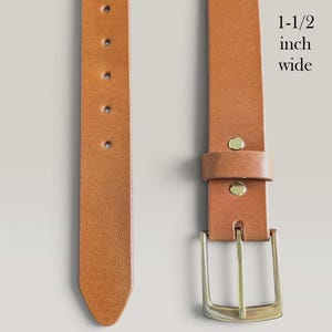 May include: A tan leather belt featuring a solid brass buckle and a series of adjustment holes. The belt is 1-1/2 inch wide and made of 9 oz full grain leather. The text 1-1/2 inch wide, 9 oz Full Grain Leather, and Solid Brass Buckle is visible.