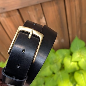 May include: A black belt featuring a smooth finish and a rectangular brass-toned metal buckle. The belt strap has a series of punched holes for adjustment and a matching black leather loop to secure the end of the strap when fastened.