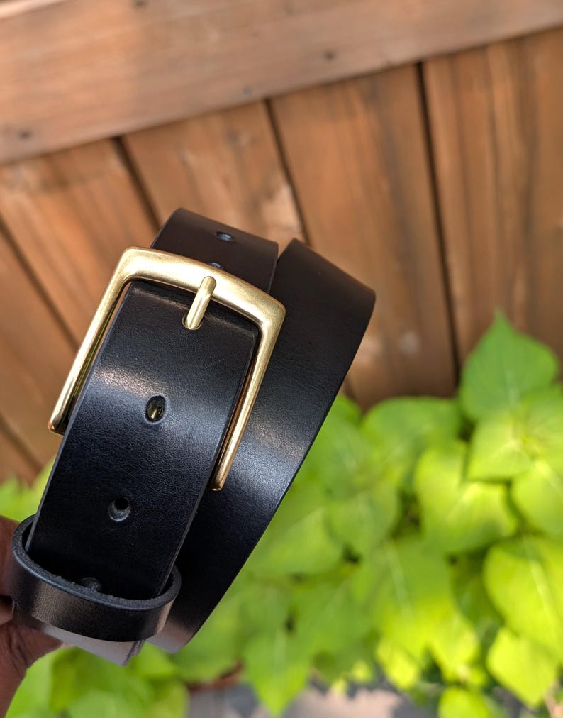 May include: A black belt featuring a smooth finish and a rectangular brass-toned metal buckle. The belt strap has a series of punched holes for adjustment and a matching black leather loop to secure the end of the strap when fastened.