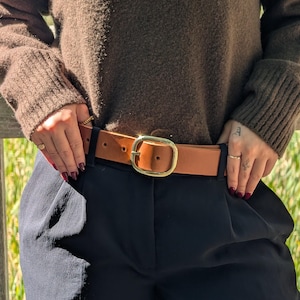 Peut inclure: Une ceinture en cuir beige avec une finition lisse et une boucle ovale dor&eacute;e polie. La ceinture est port&eacute;e dans les passants d'un pantalon noir, sous un pull &agrave; manches longues marron fonc&eacute; avec des poignets c&ocirc;tel&eacute;s et un tricot textur&eacute;.