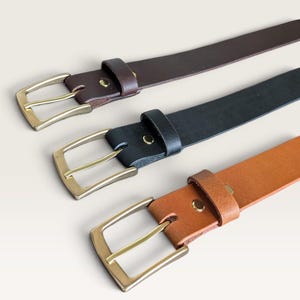 May include: Three leather belts arranged diagonally, featuring a dark brown, a black, and a tan belt. Each belt is equipped with a rectangular brass-toned metal buckle, a matching metal keeper loop, and a single metal rivet securing the strap.