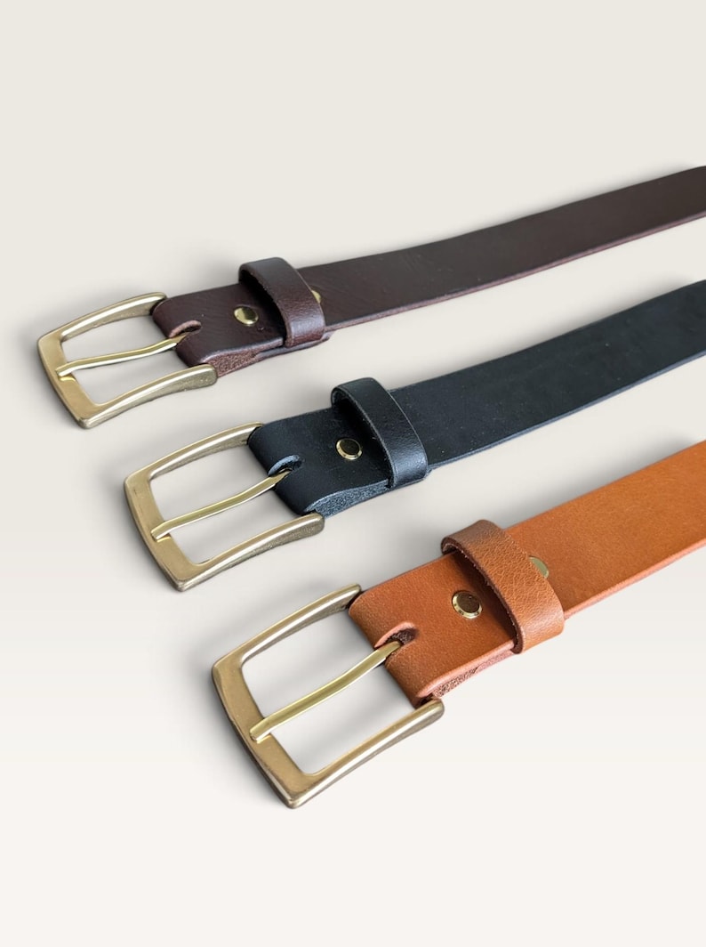 May include: Three leather belts arranged diagonally, featuring a dark brown, a black, and a tan belt. Each belt is equipped with a rectangular brass-toned metal buckle, a matching metal keeper loop, and a single metal rivet securing the strap.