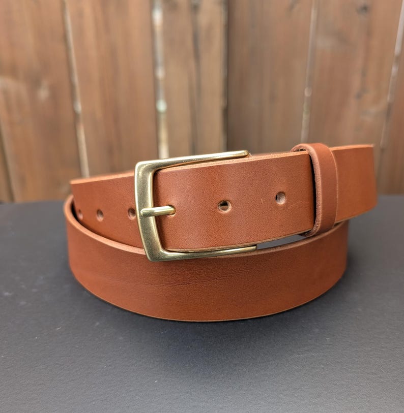 May include: A tan leather belt featuring a smooth finish and a rectangular brass-toned metal buckle. The belt includes a single leather keeper loop and multiple circular adjustment holes along the strap, coiled into a circular shape.